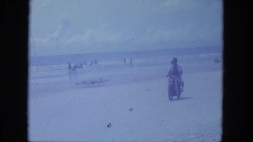 The motorcycle continues across the Clearwater beach frame, with ocean surf and scattered beachgoers behind it.