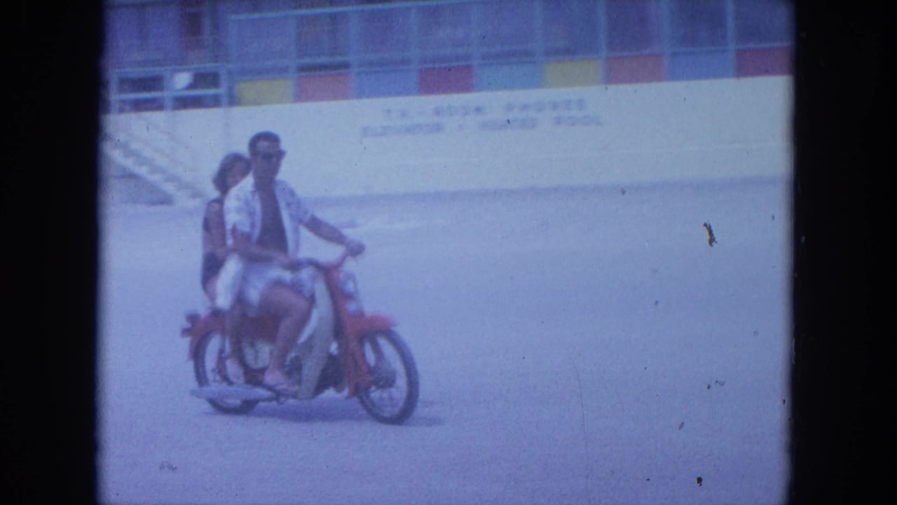 In 1964 Clearwater, Florida footage, a man in sunglasses drives a red motorcycle with a woman riding behind him near a beachside building.