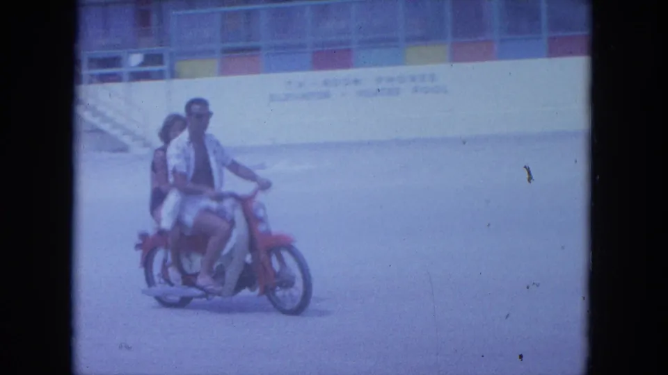 In 1964 Clearwater, Florida footage, a man in sunglasses drives a red motorcycle with a woman riding behind him near a beachside building.