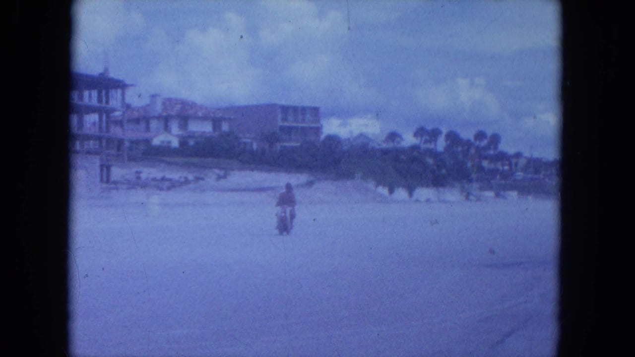 The motorcycle rides away across a broad beachside road, with low buildings, palm trees, and cloudy sky in the background.