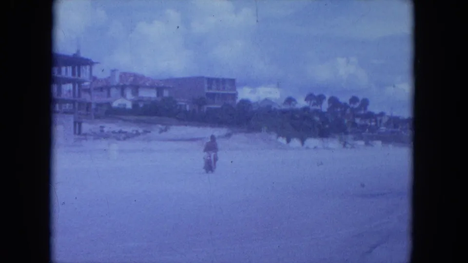 The motorcycle rides away across a broad beachside road, with low buildings, palm trees, and cloudy sky in the background.