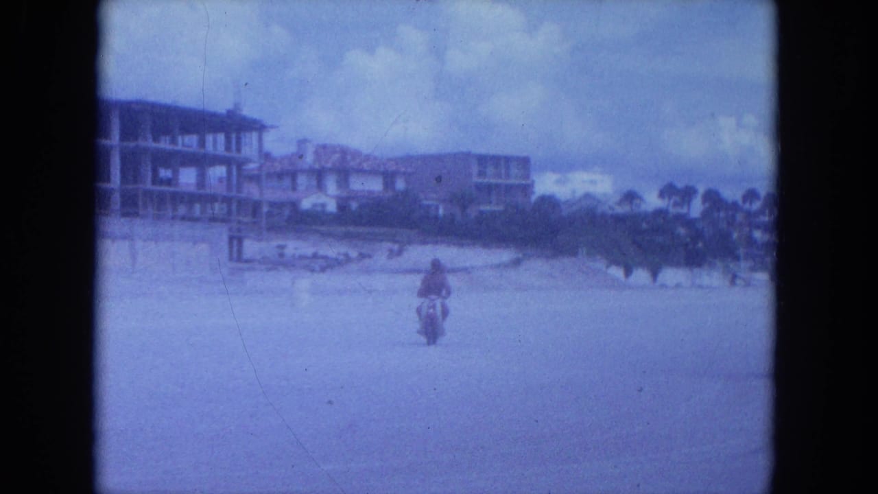 A small motorcycle figure rides away in the 1964 Clearwater beachside scene, framed by buildings, palms, cloudy sky, and 8mm film edges.
