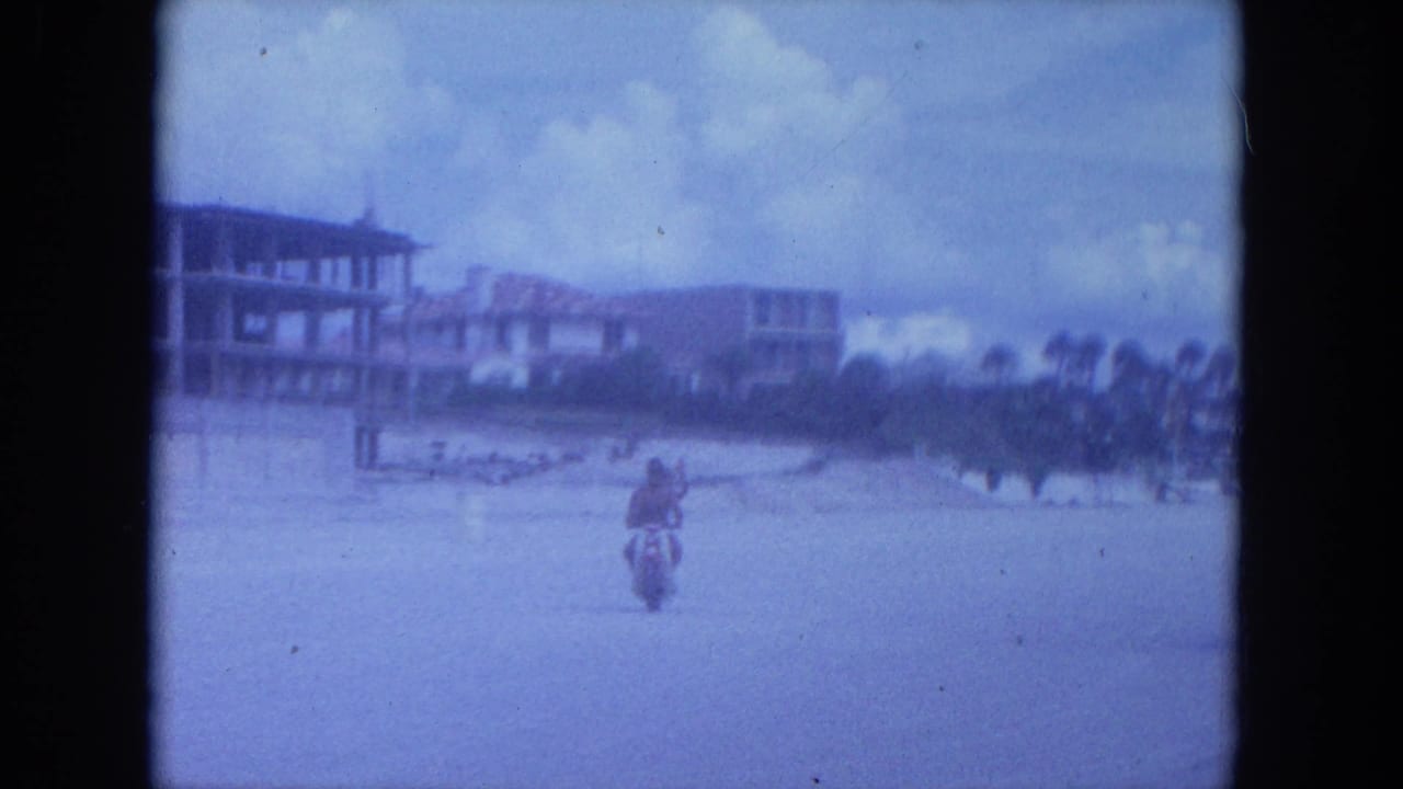 A person rides a scooter across a sandy beach with beachfront buildings in the distance.