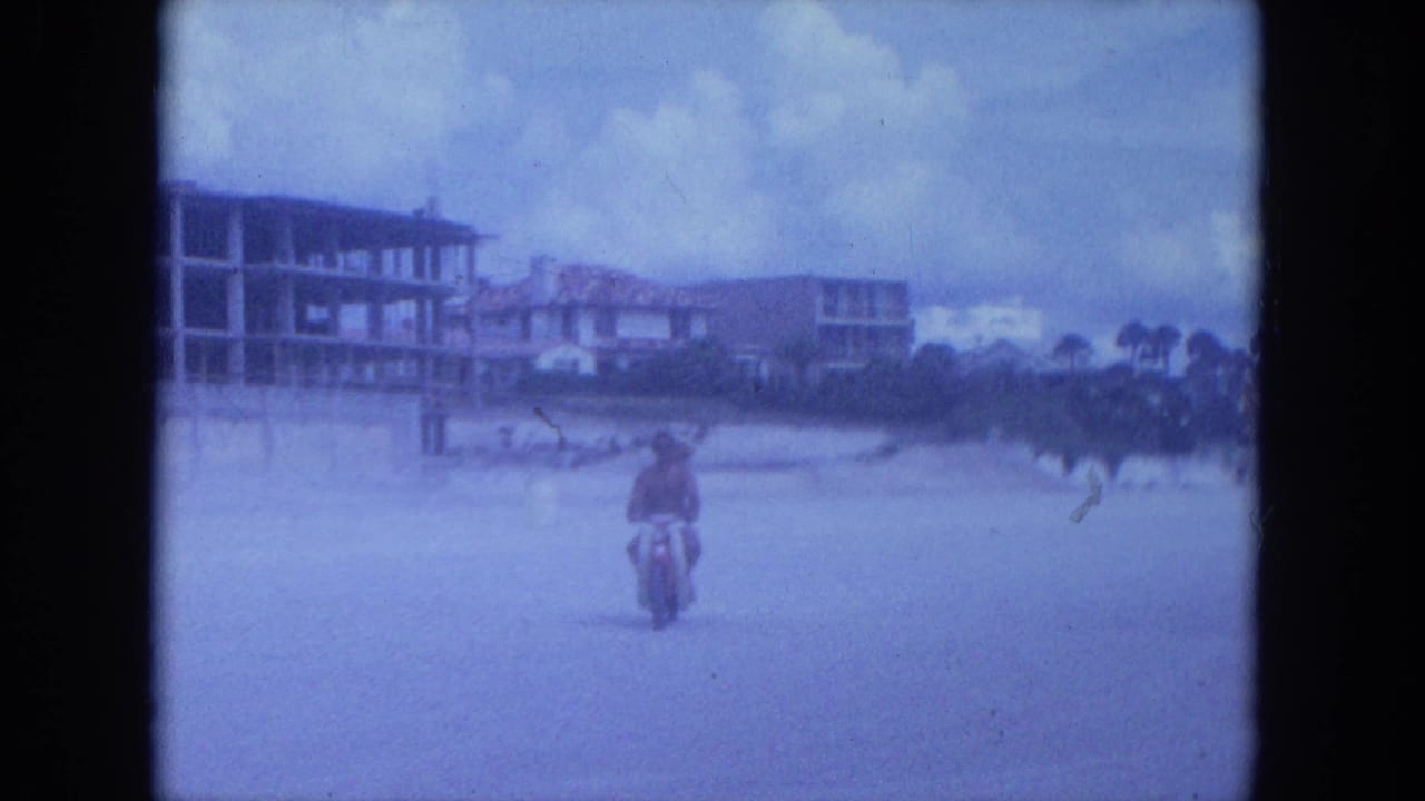 A scooter rider moves across the beach near palm trees and low buildings.