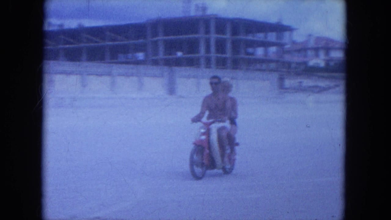 A couple rides a red scooter on Clearwater beach in 1964, passing in front of beachfront construction and soft blue 8mm sky.