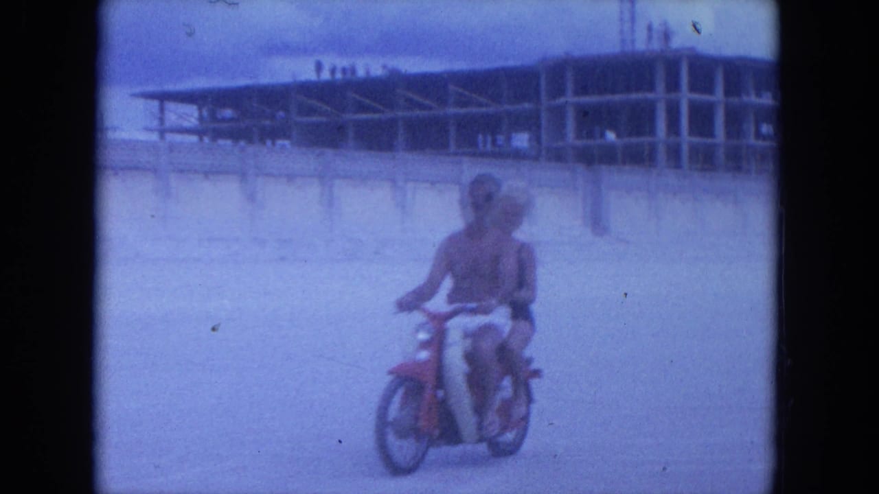 Woman rides a red scooter across sandy beach with beach buildings behind her.