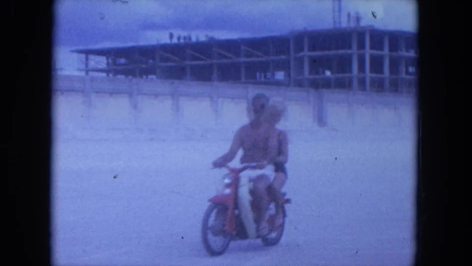 In 1964 Clearwater, Florida footage, a woman on a red scooter approaches across sand near beachside buildings.