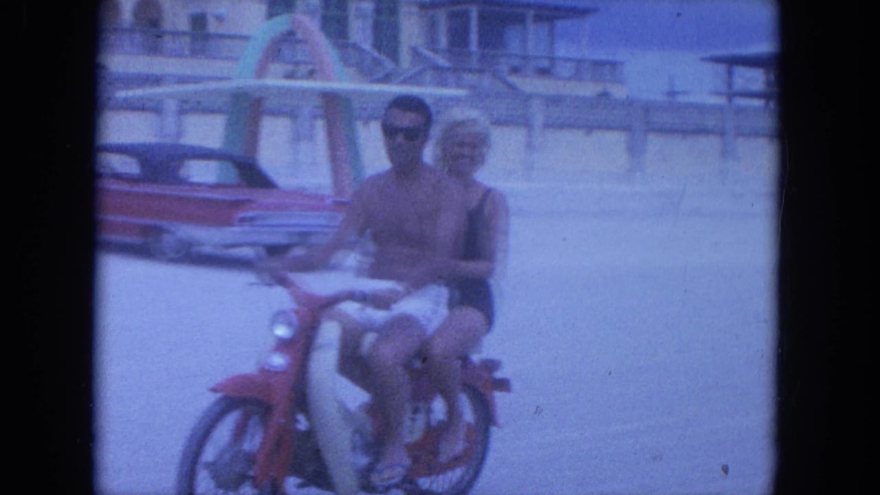 Couple rides a red scooter on the beach with a parked red car behind them.