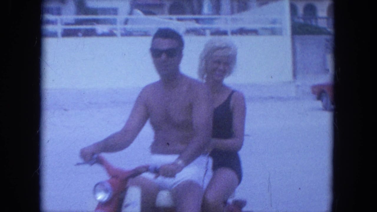 The couple rides nearer the camera on a red scooter, both in swimwear and sunglasses on the Clearwater beach.