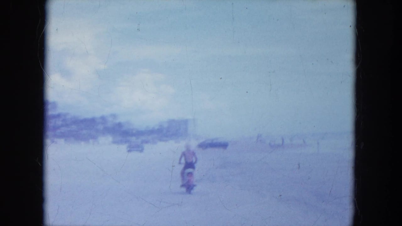 1964 Clearwater, Florida home-movie frame showing a motorcyclist on pale beach sand with distant cars and low shoreline buildings.