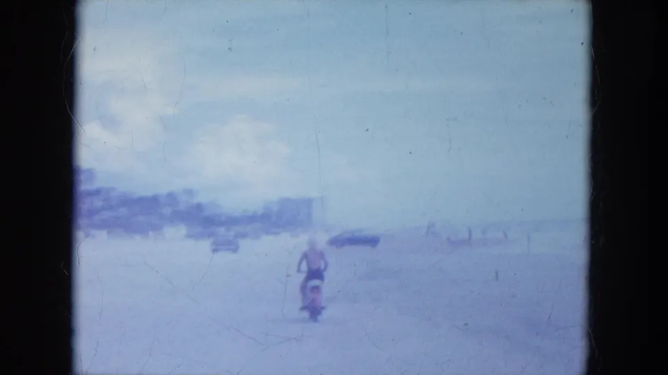 1964 Clearwater, Florida home-movie frame showing a motorcyclist on pale beach sand with distant cars and low shoreline buildings.