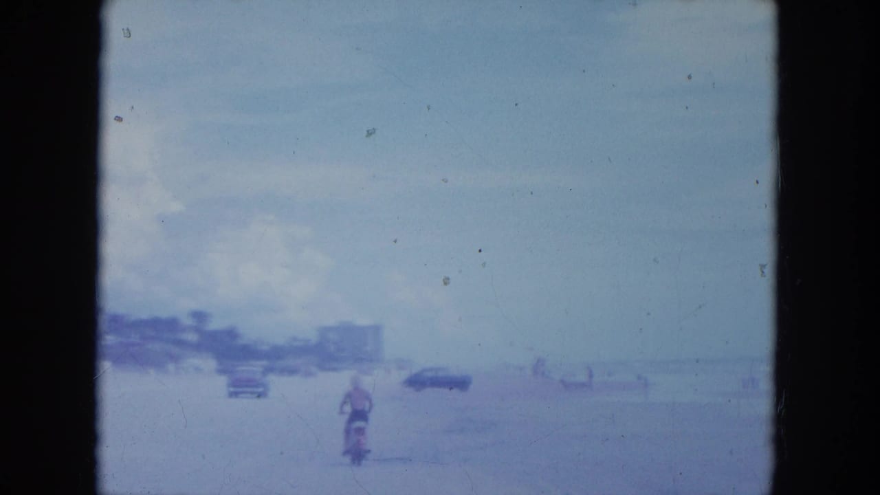 Motorcyclist continues along the beach with parked cars and shoreline figures ahead.