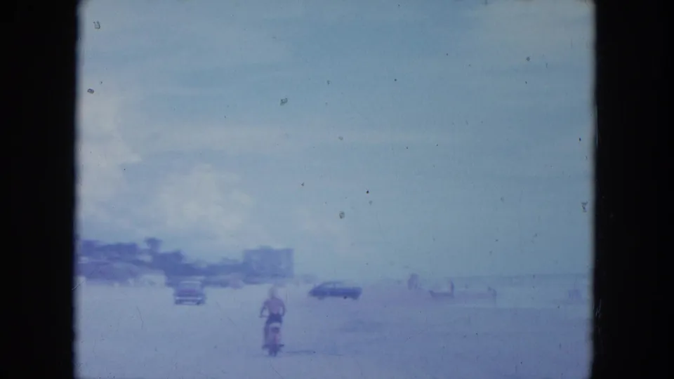 1964 Clearwater beach view with a motorcycle rider, classic cars, distant people, and a hazy shoreline seen through 8mm film grain.