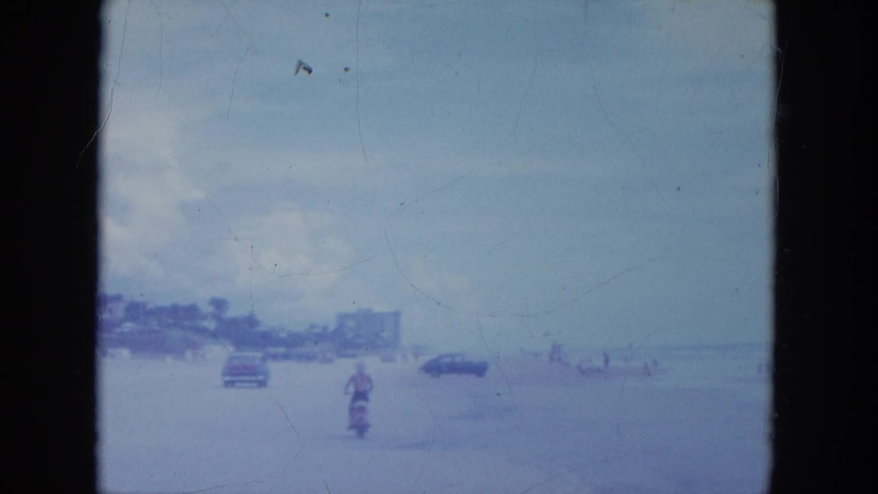 A 1964 Clearwater beach frame shows a motorcyclist near cars on open sand, with low beachfront buildings in the distance.