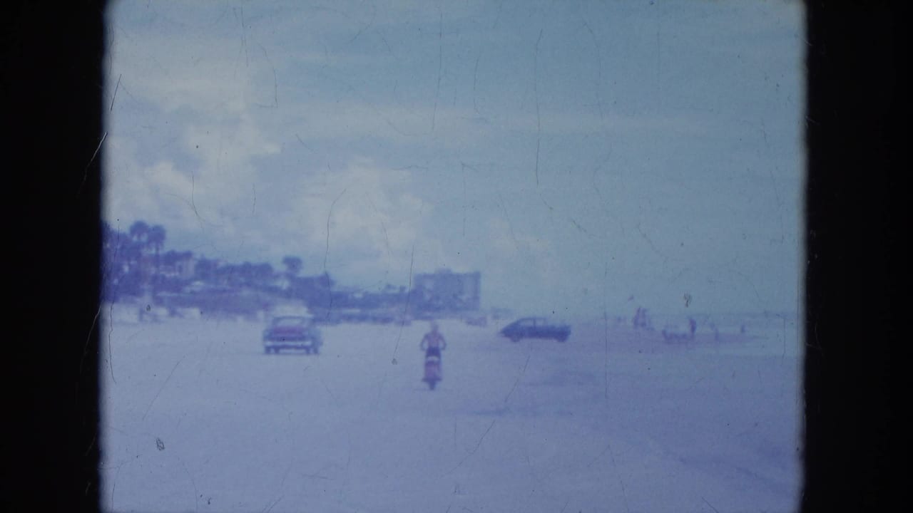 1964 Clearwater, Florida footage frame with a motorcyclist on beach sand, nearby cars, distant people, and soft 8mm exposure haze.