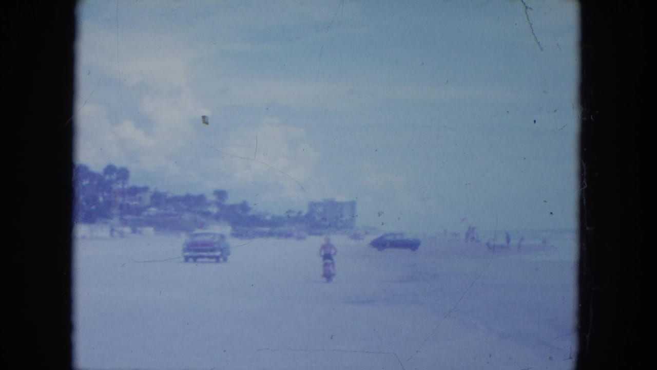 In 1964 Clearwater, a distant rider on a small motorbike crosses the beach near classic cars and low shoreline buildings.