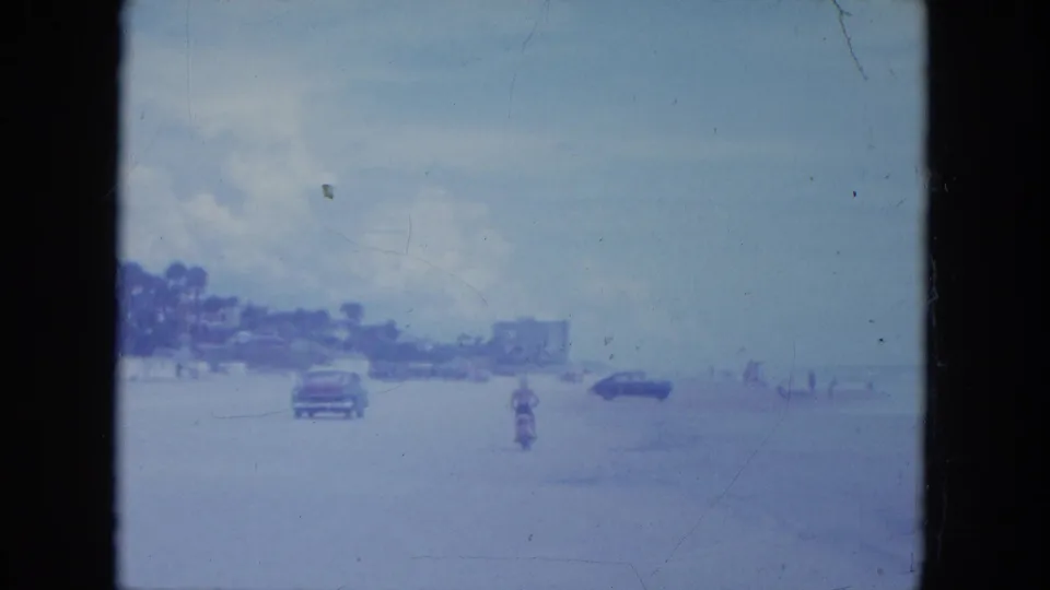 In 1964 Clearwater, a distant rider on a small motorbike crosses the beach near classic cars and low shoreline buildings.