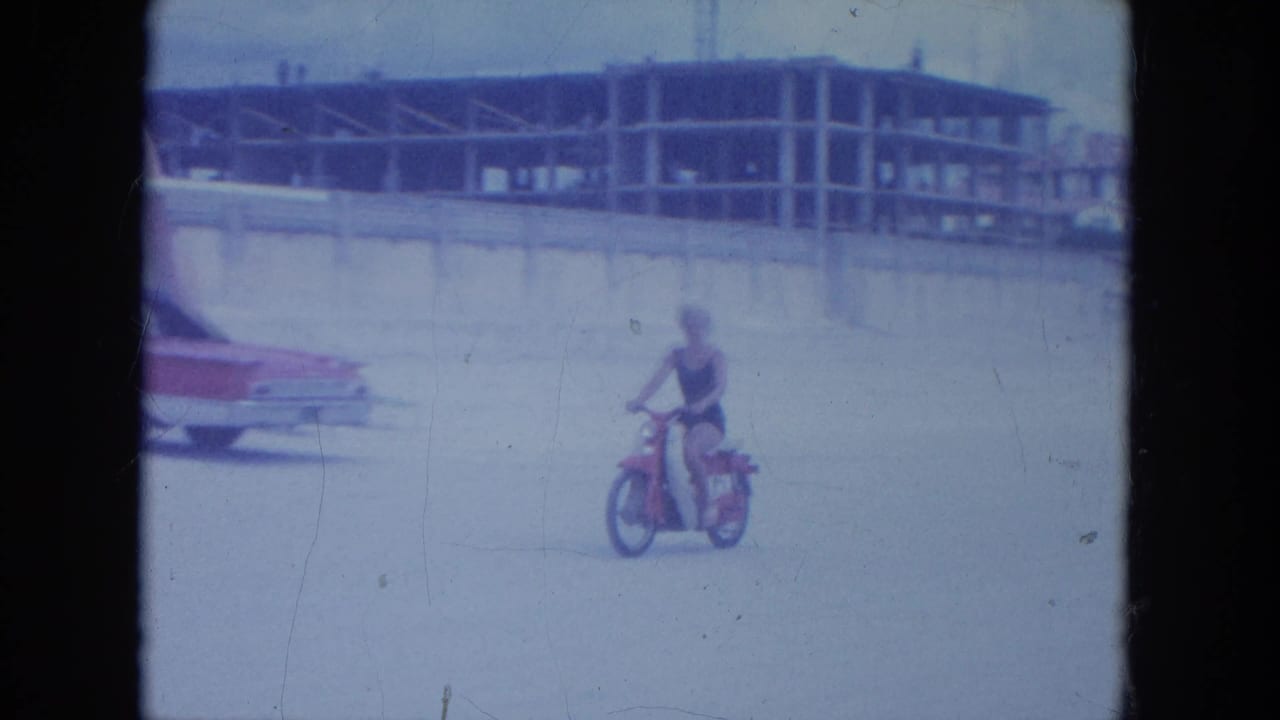 A woman in a dark swimsuit rides a red motorbike on a Clearwater beach with a classic car and unfinished building behind her.