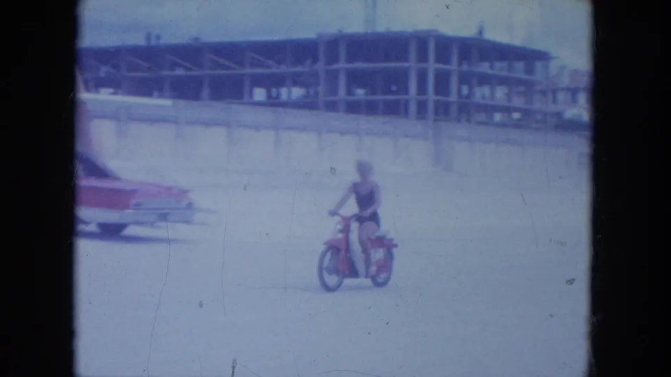 A woman in a dark swimsuit rides a red motorbike on a Clearwater beach with a classic car and unfinished building behind her.