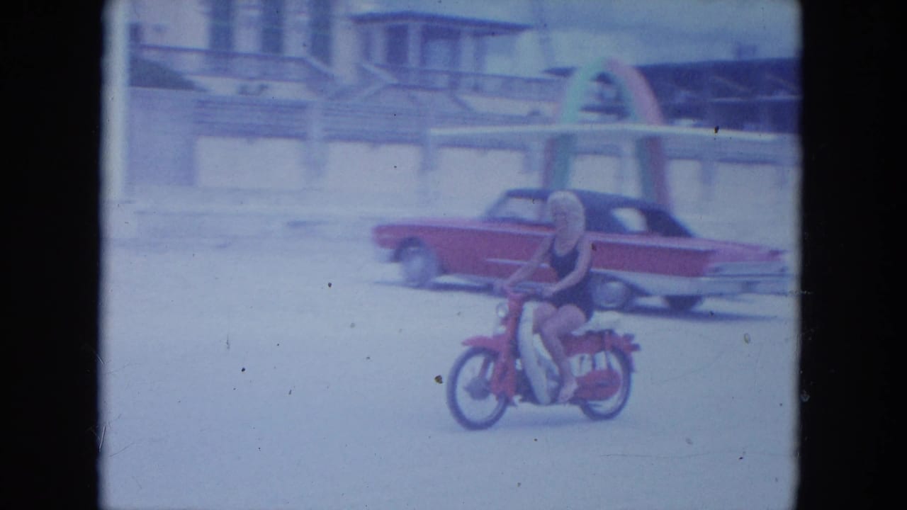 The woman rides the red motorbike across the sand, framed closer beside a red and white classic American car.