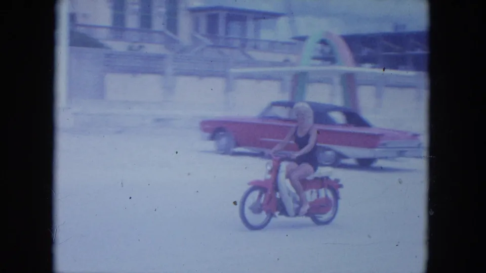 The woman rides the red motorbike across the sand, framed closer beside a red and white classic American car.
