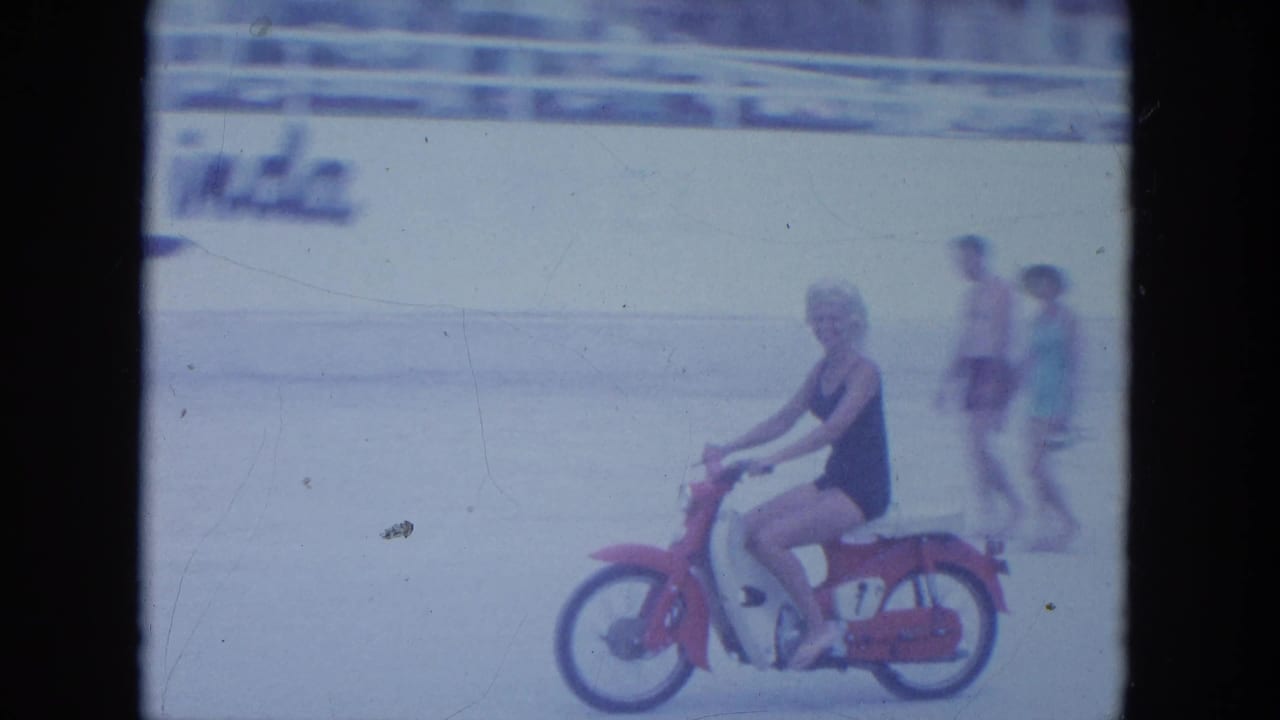 The rider continues along the Clearwater beach while two pedestrians pass near a wall with visible lettering in the background.