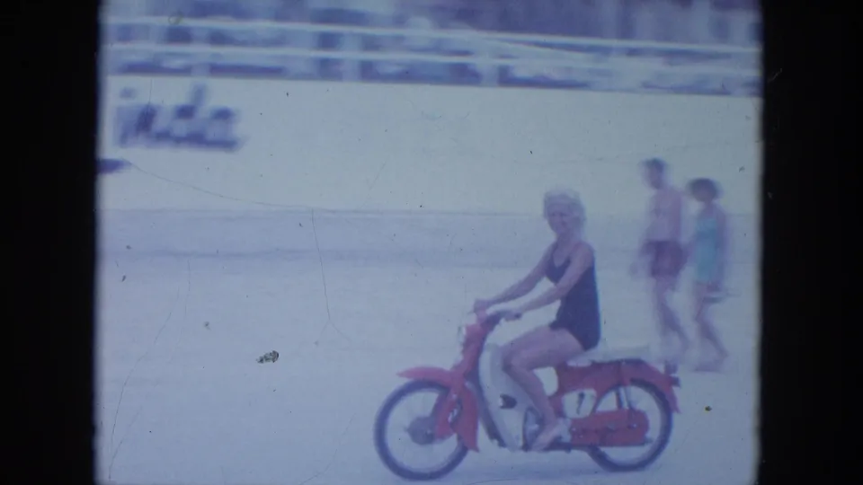 The rider continues along the Clearwater beach while two pedestrians pass near a wall with visible lettering in the background.