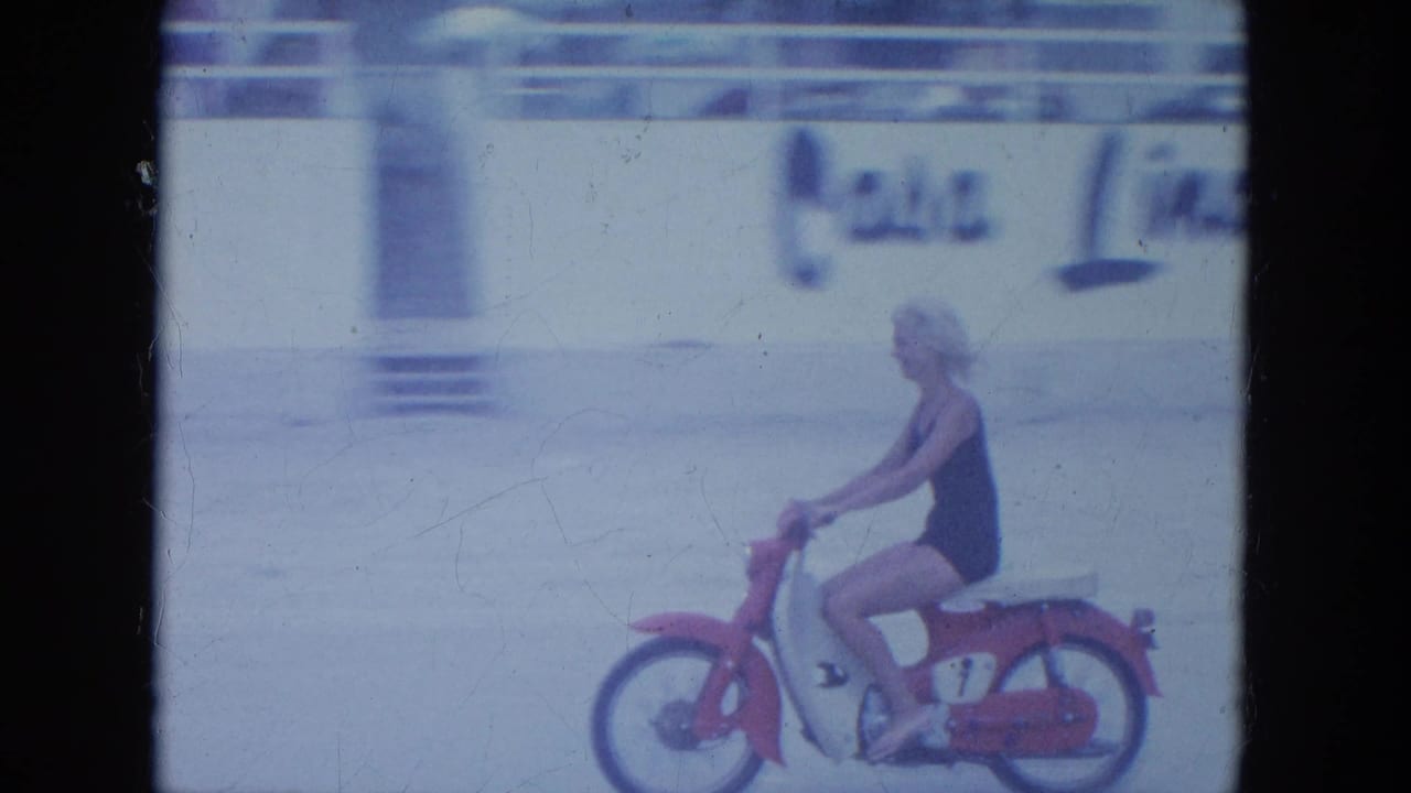 Woman in a dark swimsuit rides a red-and-white motorbike across pale beach sand.