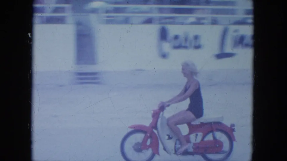 In Clearwater, Florida, 1964, an older woman in a dark one-piece swimsuit rides a red-and-white motorbike past beachside buildings.