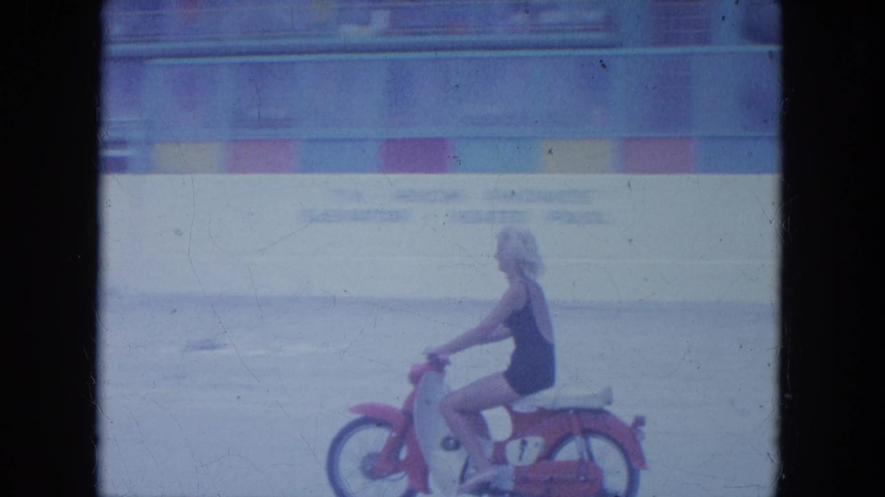 Woman on a small red motorbike continues across the beach near a low wall.