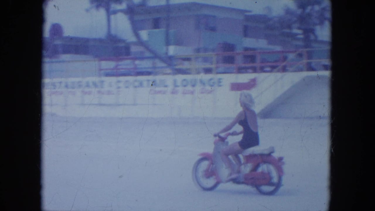 The woman rides past beach frontage with a railing and partially legible lounge signage in the 1964 Clearwater home-movie footage.