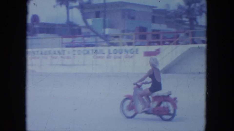 The woman rides past beach frontage with a railing and partially legible lounge signage in the 1964 Clearwater home-movie footage.