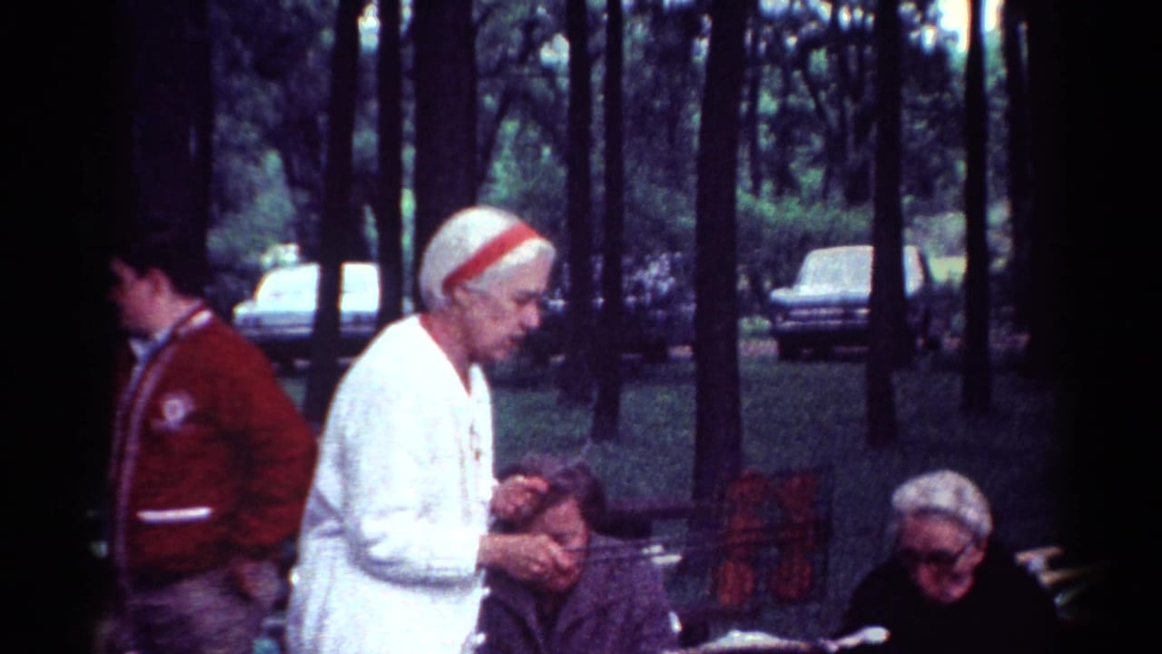 Priest in white stands near seated adults in a wooded park.