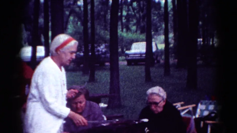 Priest in white stands beside seated women during a 1964 Cleveland park gathering, with cars visible through trees.