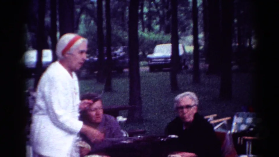 1964 Cleveland home-movie frame with a priest in white standing beside seated adults at a wooded park table.