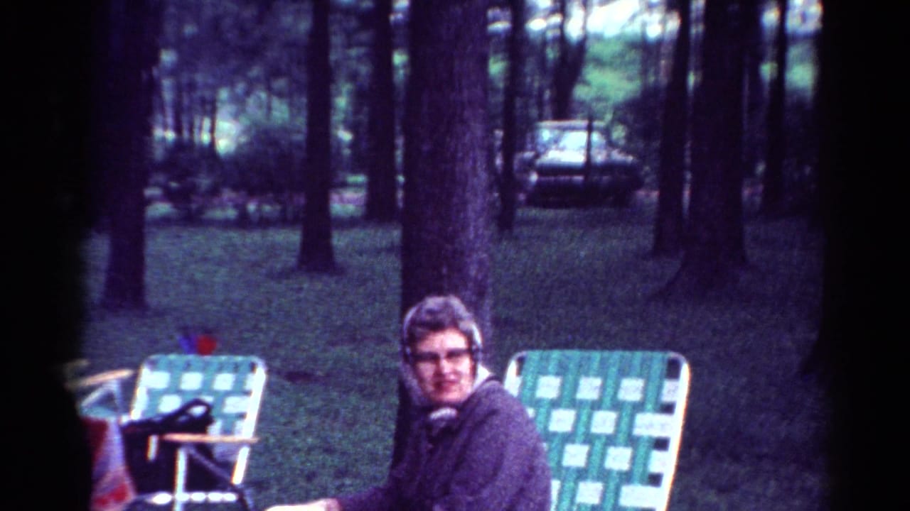 1964 Cleveland home-movie frame showing a woman in glasses seated in a checkered lawn chair in a wooded picnic area.