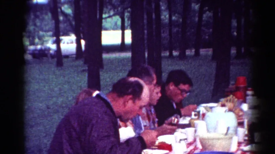 1964 Cleveland home-movie frame showing adult men gathered at a picnic table during an outdoor meal.