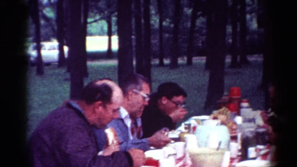 1964 Cleveland park picnic frame with adult men eating at a crowded table in a shaded wooded area.