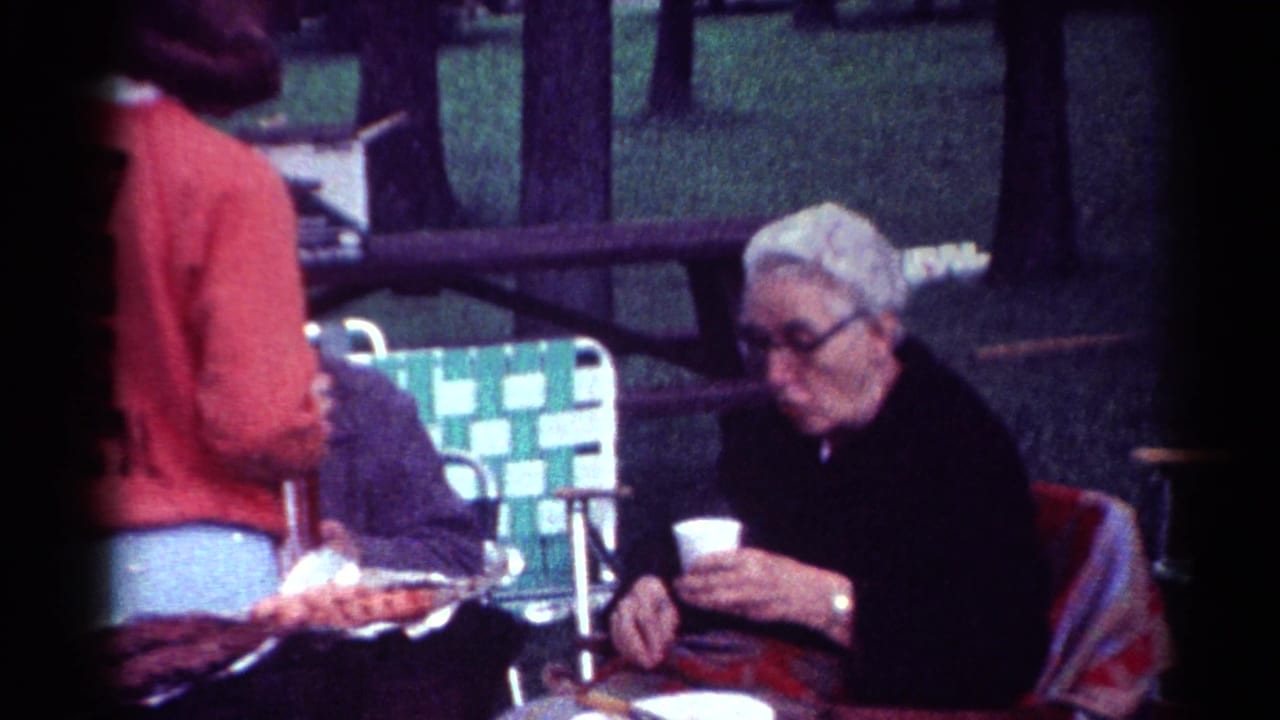 Older man with white hair and glasses sits at a picnic table holding a cup while a woman stands nearby.