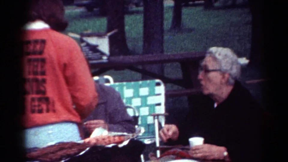 1964 Cleveland picnic scene with a red slogan sweatshirt, seated older man, paper cup, folding chair, and wooded park background.