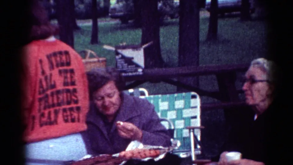 1964 Cleveland park picnic frame showing a person in a purple jacket eating beside an older man and a red sweatshirt in the foreground.