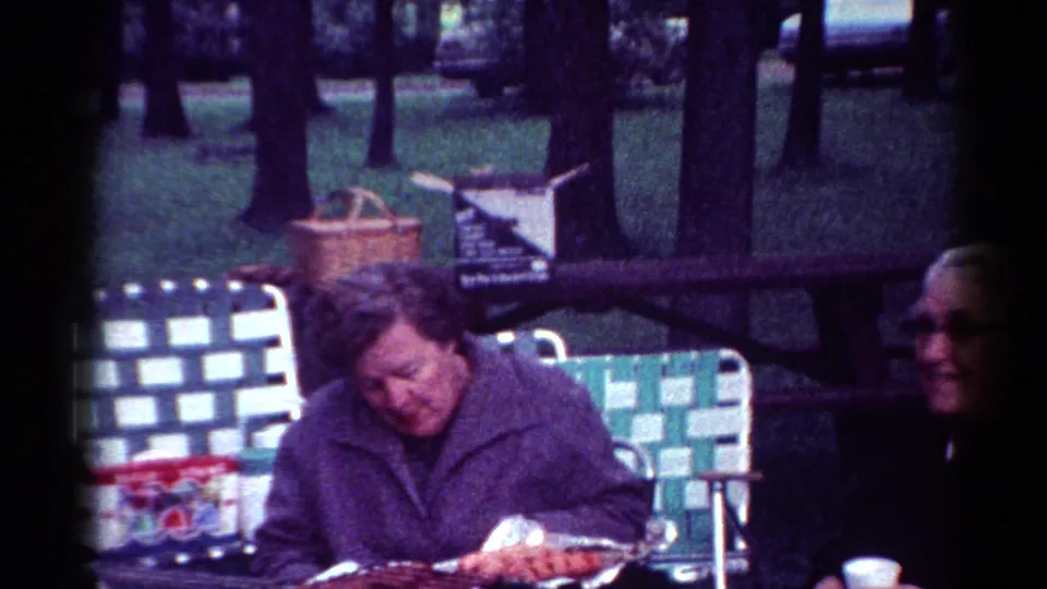 1964 Cleveland park picnic frame with a purple-jacketed guest looking down, a paper cup, woven chair, trees, and an older man at the edge.