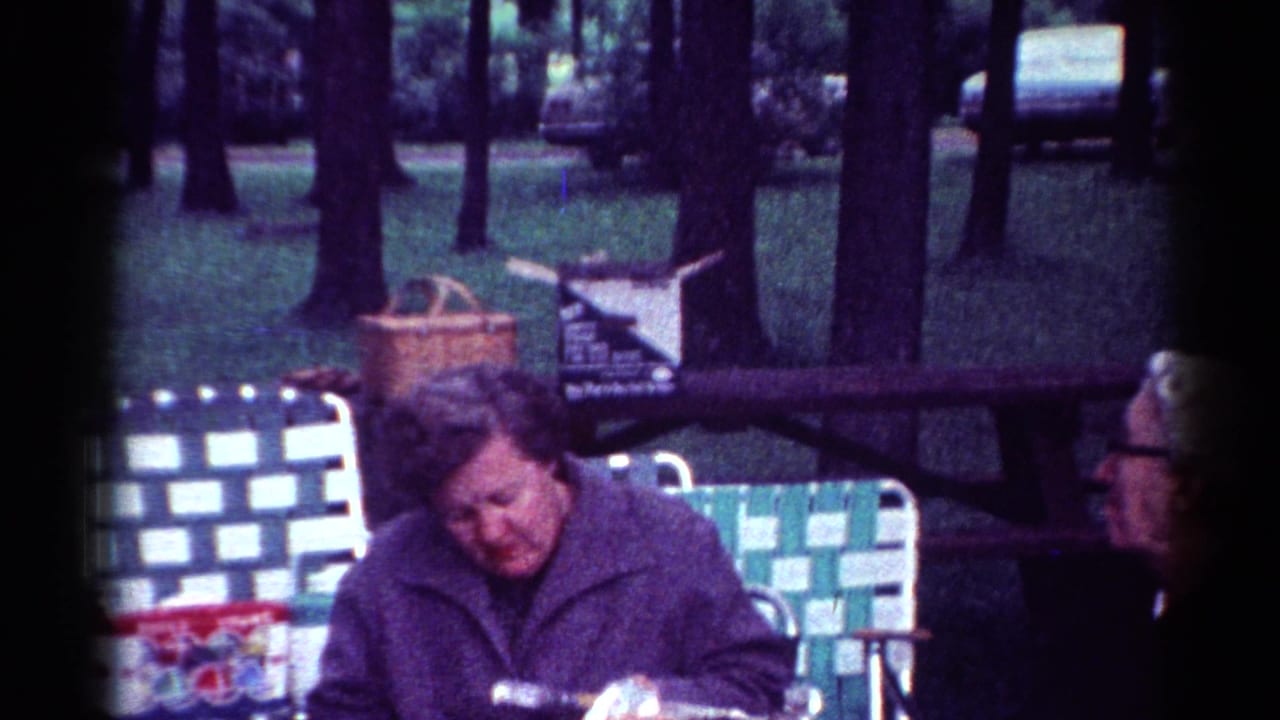 Woman seated at a picnic table with lawn chairs and trees behind her.