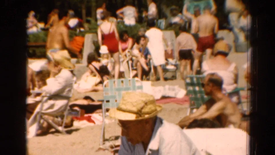 1964 Cleveland home-movie beach frame showing a dense crowd of adults in swimsuits, beach chairs, towels, and sunlit sand.