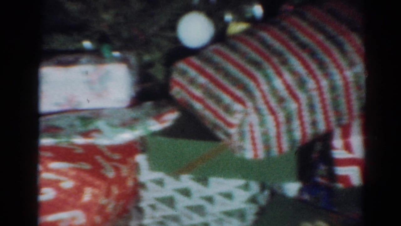 1964 Cleveland home-movie frame showing wrapped Christmas presents in red, white, and green patterned paper inside a dim room.