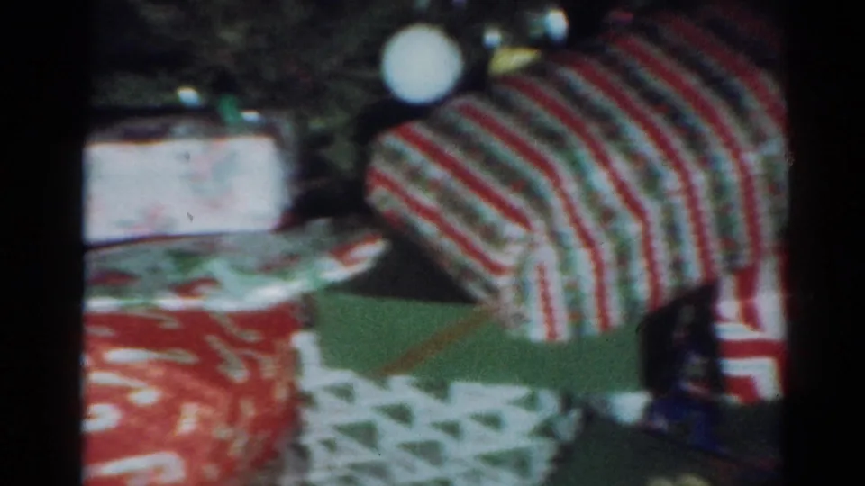 1964 Cleveland home-movie frame showing wrapped Christmas presents in red, white, and green patterned paper inside a dim room.