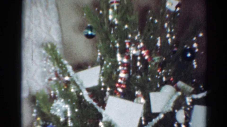1964 Cleveland home-movie frame showing a close Christmas tree detail with silver tinsel, colored ornaments, striped garland, and small white cards.