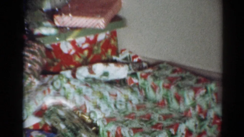 1964 Cleveland frame showing wrapped Christmas presents in patterned paper stacked near the wall with partial holiday decorations above.