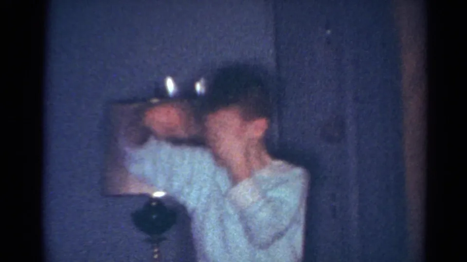 1964 Cleveland home-movie frame showing a young person in a light sweater reacting beside a lit candle in a private interior.