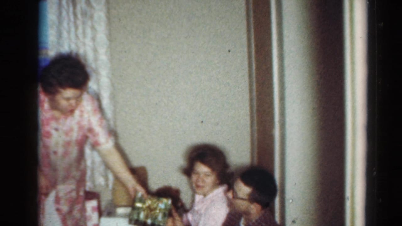 1964 Cleveland home-movie frame of a woman standing by seated relatives in a living room with wrapped gifts nearby.